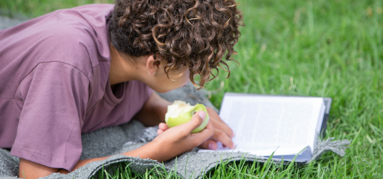 Ungdom med äpple i handen ligger på en filt och läser en bok