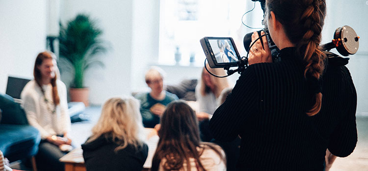 En grupp vuxna runt ett bord blir filmade av en kvinna med stor filmkamera